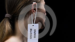 Male hand hanging dumb blond label on female ear, social prejudice and sexism