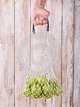 Male hand hold juicy organic green apples in a cotton mesh bag on a wooden table. Vertical shot