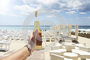 Male hand holding a bottle of beer against a sunny sky and crystal clear sea. In the background a beautiful beach resort. Vacation