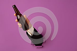 Male hand holding bottle of beer breaks through purple background