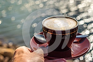 Male hand holding a ceramic cup of cappuccino under the sun, outdoors