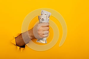 Male hand holding crumpled dollar banknotes through hole in yellow background with copy space