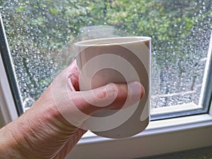 Male hand holding a cup of coffee or tea on a rainy day window background