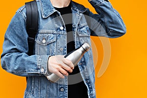 Male hand holding eco steel thermo bottle for water, wearing jeans jacket and backpack, on yellow background.