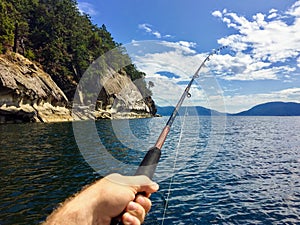 A male hand holding a fishing rod jigging for fish with the beautiful coastline of British Columbia Canada in the background.