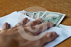 Male hand holding its fingers on white envelope full of American Dollars (USD, US Dollars) on the wooden table as a symbol of