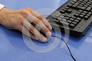 Male hand is holding a mouse next to the keyboard. Close-up. An office worker or manager works on a computer. Blue background