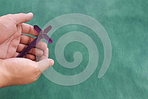 Male hand holding purple wooden cross flat lay in green background. Holy week and lent season celebration concept