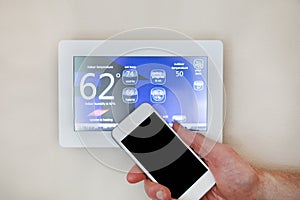 Male hand holding smart phone to operate heating or cooling via digital touch screen thermostat for home photo