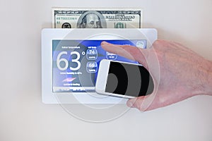 Male hand holding smart phone to operate heating or cooling via digital touch screen thermostat for home energy savings