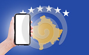 Male hand holding smartphone with blank on screen, on background of blurred flag of Kosova. Close-up view.