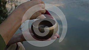 Male hand holding Tibetan singing bowl and making sounds out of it against river background