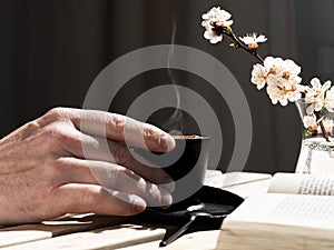 Male hand holds a cup of coffee with aromatic espresso. Wooden vintage table. Next to the open book and a branch of blooming