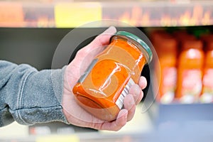 Male hand holds jar of sauce on blurred background, row of shelves with groceries in supermarket, concept of marketing, prices for