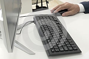Male hand holds a mouse next to the keyboard and monitor of a personal computer on a white table. Close-up. A businessman, clerk,