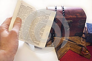 The male hand holds a new testament on a white background. Alongside there is a casket with a variety of jewels. The gold chain is