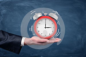 A male hand holds palm up a large red alarm clock with white sound wave lines on blue background.