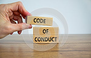 Male hand placing a block with word `code` on top of a blocks with words `code of conduct`. Beautiful wooden table, white