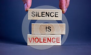 Male hand placing a block with word `silence` on top of a blocks tower with words `silence is violence`. Beautiful grey backgr