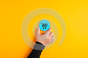 Male hand placing a blue badge with a shopping cart icon on yellow background. Shopping or online e-commerce