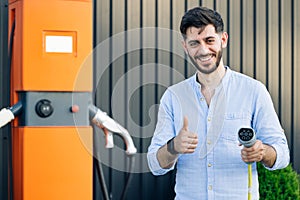 Male hand showing thumbs up holding power cable supply plugged at electric car charging station. Bearded man standing