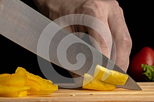 Male hand slicing yellow bell pepper with sharp metal knife. Cook grinds pulp of bell pepper on wooden cutting board