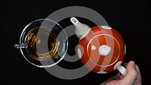 Male hand takes a cup of tea from a transparent glass next to a red and white polka dot teapot on a black background 4k