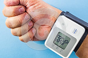 Male hand wearing white and grey wrist blood pressure monitor on blue background