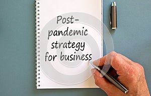 Male hand writing `post-pandemic strategy for business` on white note,  on blue background. Business concept