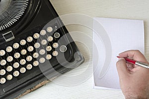 A male hand writing on a white checkered paper with a red pen next to an old wrecked black typewriter