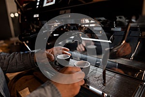 Male hands baristas hold two cups near the modern coffee machine. Professional coffee machine pours hot coffee drink in a mug.