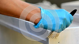 Male hands in blue protective gloves hold a paintbrush and paint a new wooden plank with white paint outdoors. The man
