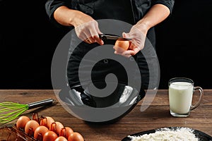 Male hands chef chefs break the egg on a wooden brown table in a black bowl. Black background. The concept of cooking food. Close-