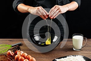 Male hands chef chefs break the egg on a wooden brown table in a black bowl. Black background. The concept of cooking food. Close-