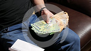 Male hands are counting fifty and one hundred euro banknotes on a leather black sofa in the room. Money in the hands and