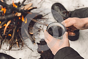 Male hands hold a mug of coffee near a burning campfire. Concept hike, walk, trip in winter