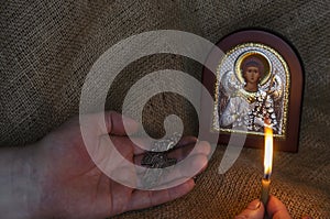 Male Hands hold a pectoral cross and a burning candle in front of a Christian icon