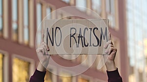 Male hands hold a poster with the slogan NO RACISM against the background of the city. Protest poster close up outdoors