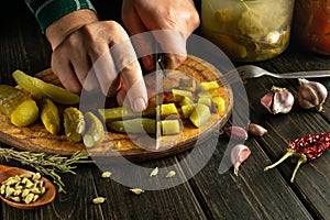 Male hands with a knife cut pickled cucumber into small pieces on a kitchen board to prepare a salad for breakfast