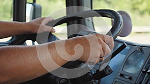 Male hands of lorry driver holds a big steering wheel while driving a truck at summer day. Trucker riding to destination