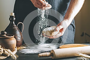Male hands pour flour on the dough next to clay pot and oil bottle and rolling pin on dark table, while cooking