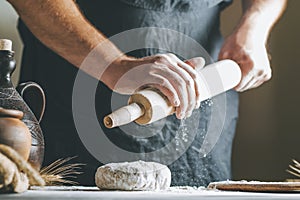 Male hands pour flour on a rolling pin next to dough, clay pot, oil bottle and rolling pin on dark table, while cooking