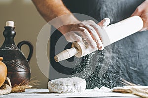 Male hands pour flour on a rolling pin next to dough, clay pot, oil bottle and rolling pin on dark table, while cooking