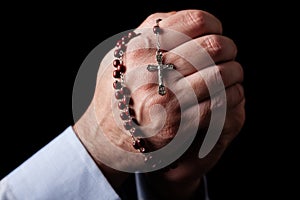 Male hands praying holding a rosary with Jesus Christ in the cross or Crucifix on black background.