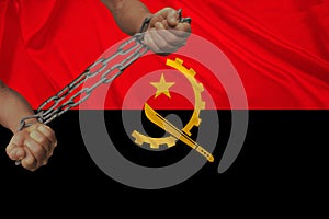 Male hands of a prisoner in iron chains against the background of the national flag of Angola on a beautiful silk fabric, the