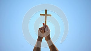 Male hands raising wooden cross to blue sky, religious conversion, baptism