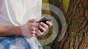 Male hands with a smartphone, close-up. Writes text against the backdrop of a large tree and countryside