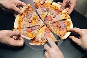 Male hands taking slices of pizza with cheese, tomatoes and ham from food delivery. Group of hungry friends sitting at