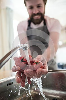 Male hands wash red tomatoes under running water in a sink. Smiling man in an apron is preparing for cooking, in the