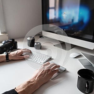 Male hands working at the computer on a white desktop with photographic equipment and coffee. Man photographer freelancer works.